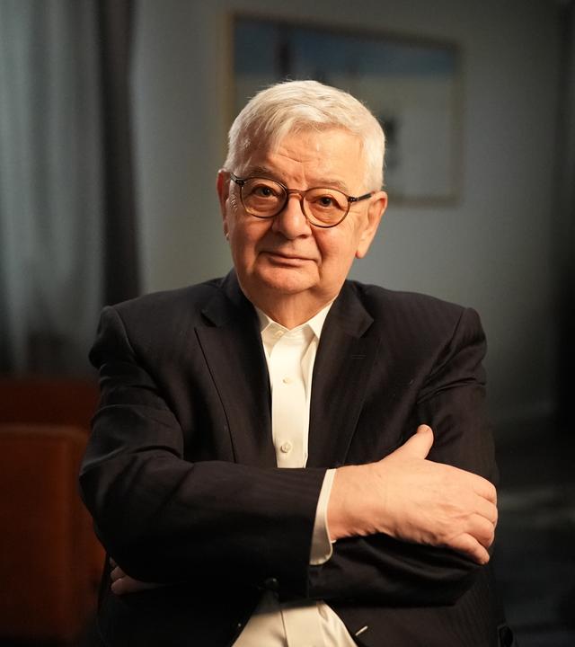 Joschka Fischer sitzt in einem dunklen Raum und schaut in Richtung Kamera.