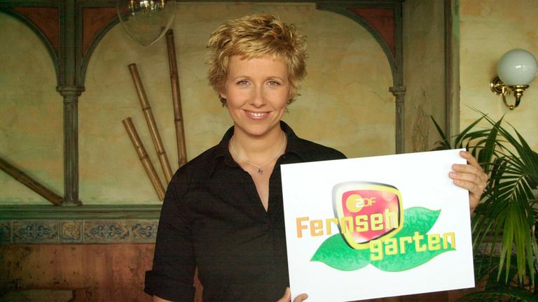 Andrea Kiewel hält das Fernsehgarten Schild in der Hand