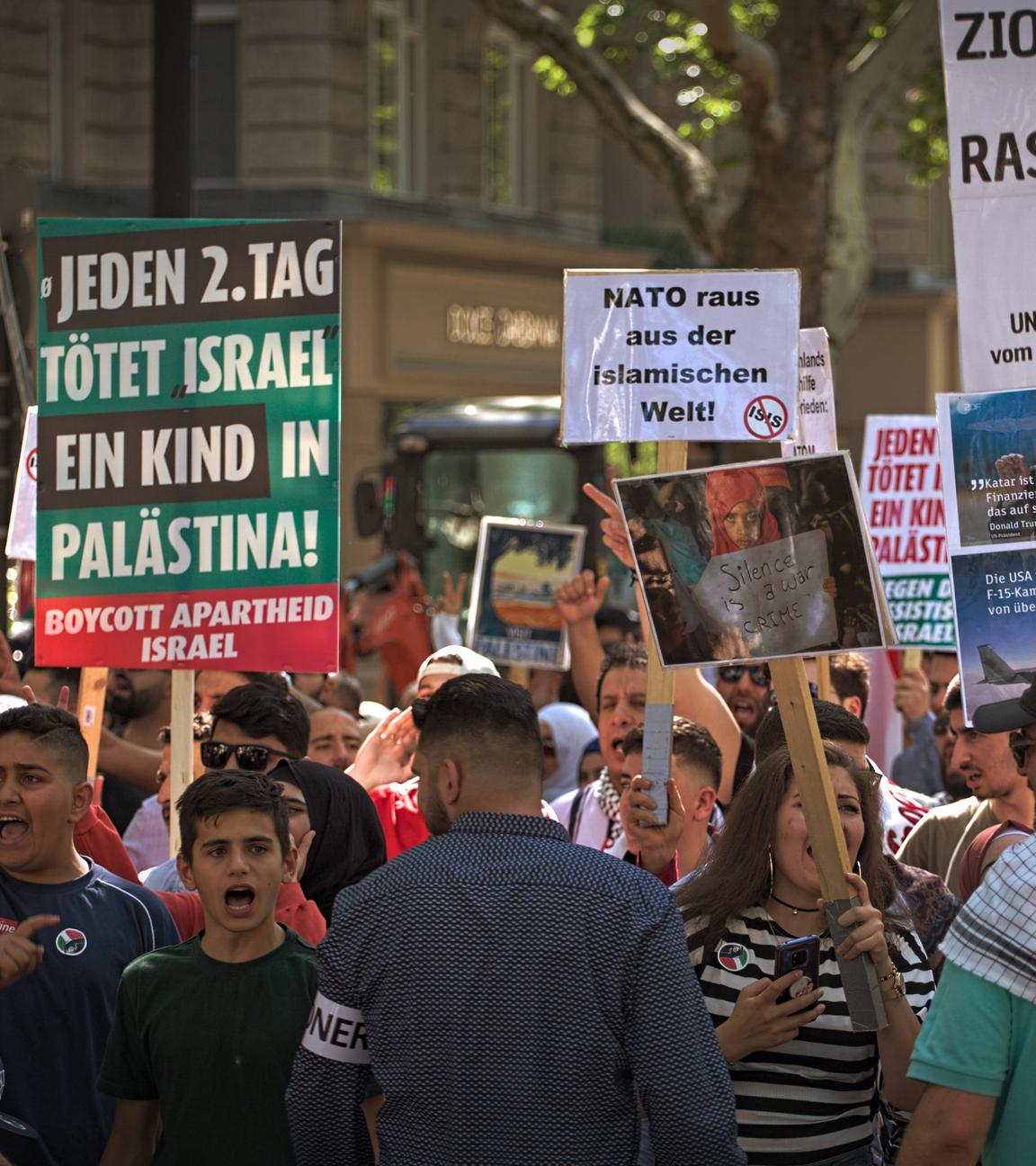 Während einer Demonstration halten mehrere Menschen Schilder in die Luft und schreien. Auf einem der Schilder steht die Aufschrift "Jeden 2. Tag tötet Israel ein Kind in Palästina".