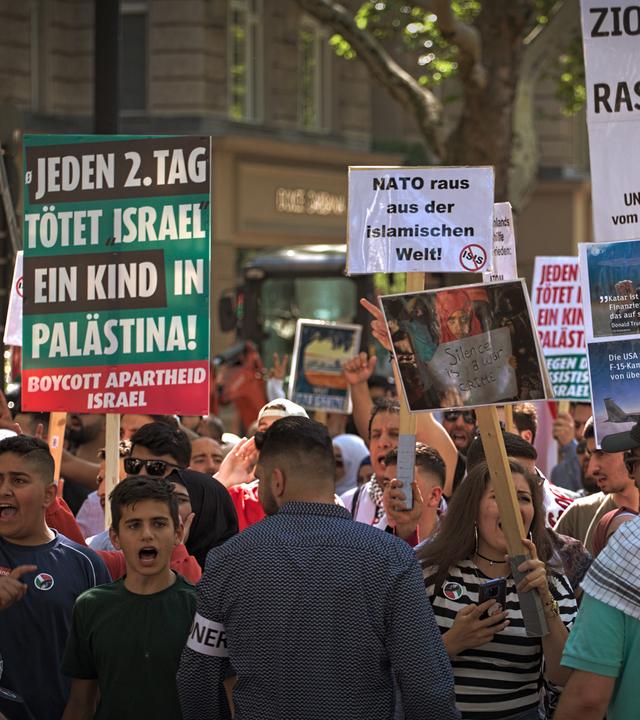 Während einer Demonstration halten mehrere Menschen Schilder in die Luft und schreien. Auf einem der Schilder steht die Aufschrift "Jeden 2. Tag tötet Israel ein Kind in Palästina".