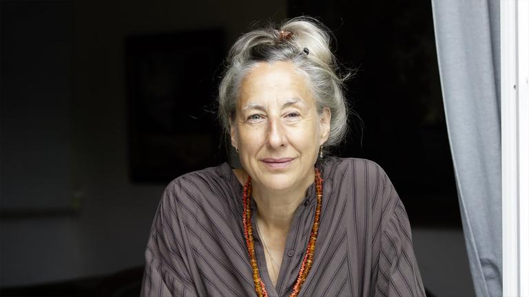 Porträtfoto einer Frau mittleren Alters mit hochgesteckten graublonden Haaren, die eine lange doppelreihige orangefarbene Edelsteinkette trägt.