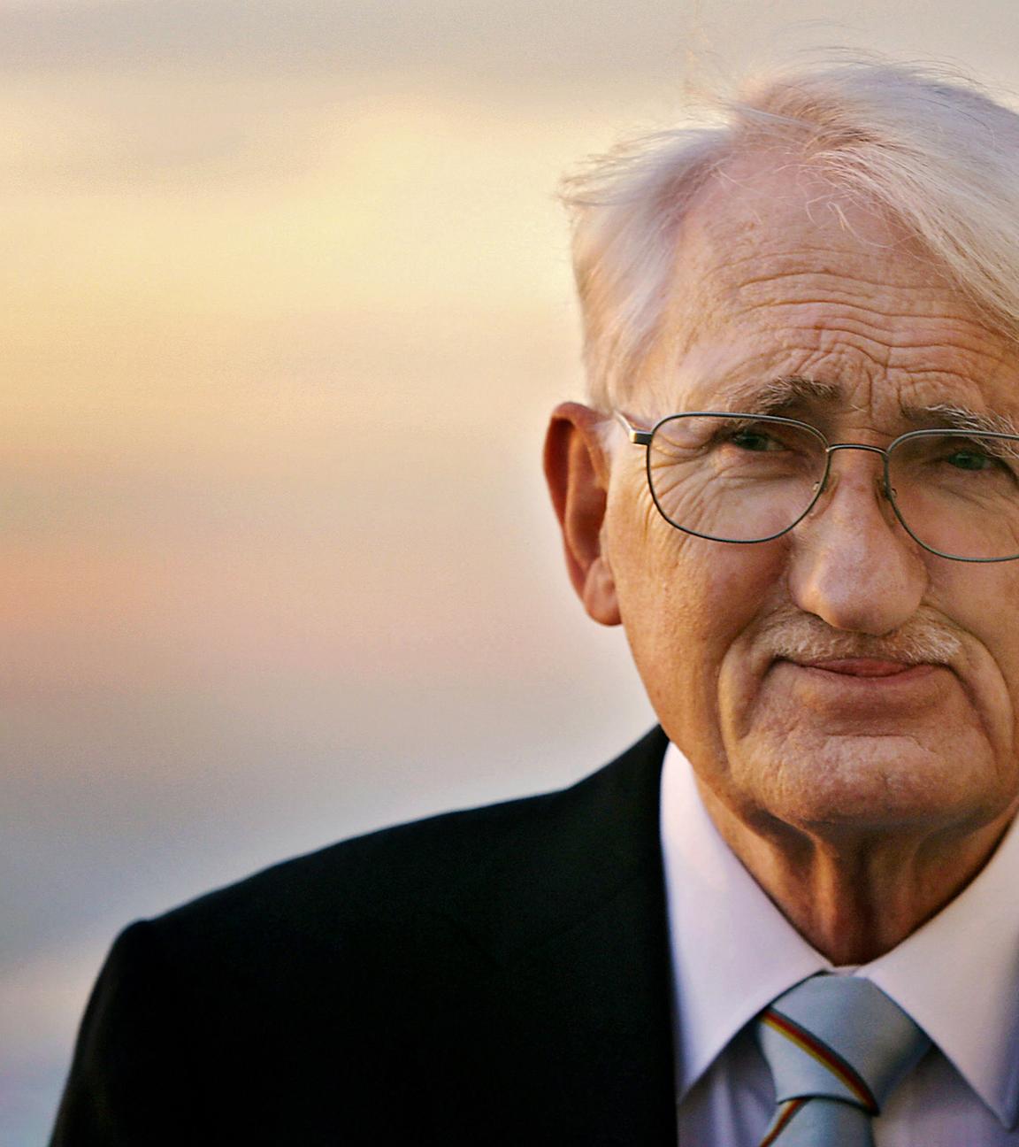 Porträtfoto eines älteren Mannes mit kurzen weißen Haaren und Schnurrbärtchen, der eine Brille und einen Anzug trägt und vor einem Sonnenuntergang steht.