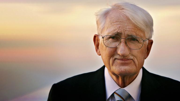 Porträtfoto eines älteren Mannes mit kurzen weißen Haaren und Schnurrbärtchen, der eine Brille und einen Anzug trägt und vor einem Sonnenuntergang steht.