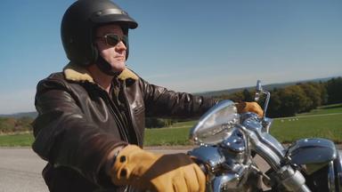 Auf dem Bild ist zu sehen: Jürgen Tonkel mit schwarzem Helm, Sonnenbrille und Lederjacke auf einem Motorrad.