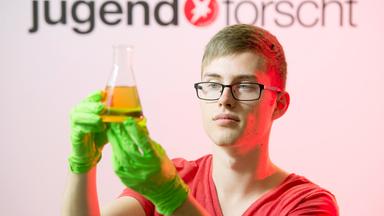 Ein Schüler vor dem Logo des Wettbewerbs "Jugend forscht" mit einem Reagenzglas.
