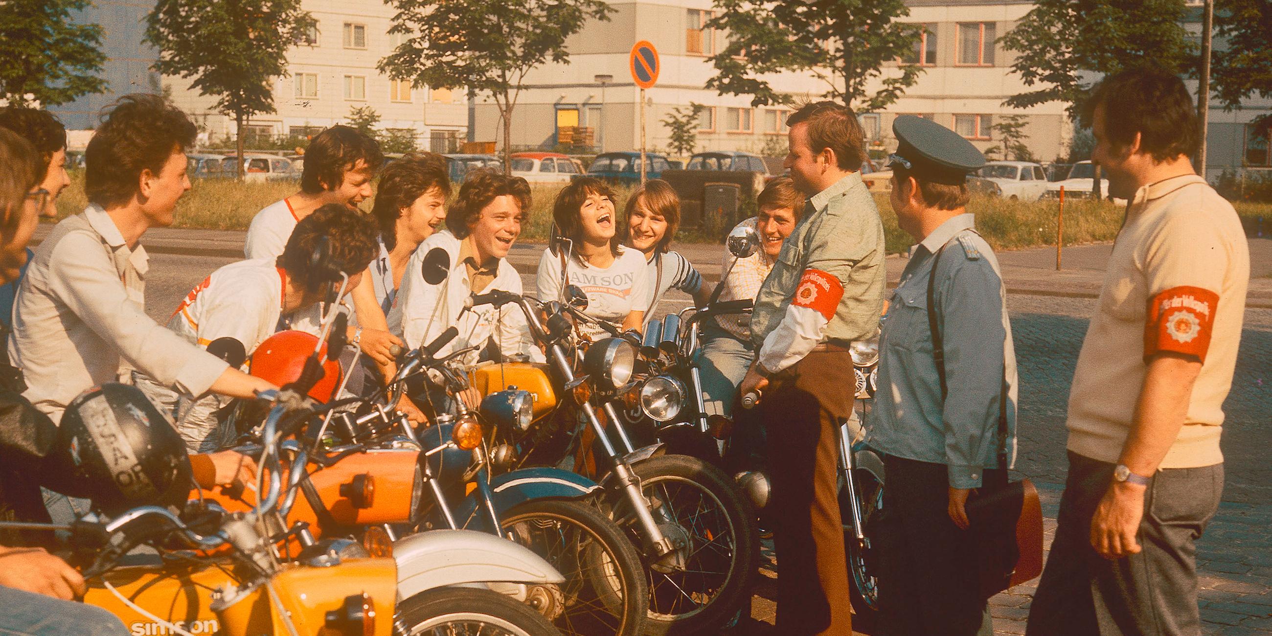  Mehrere Jugendliche sitzen an einer Straße auf ihren Mopeds und sprechen mit drei DDR-Volkspolizisten.