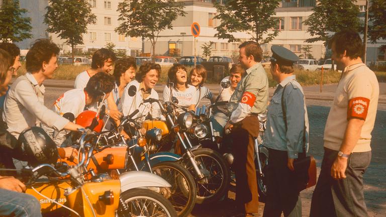  Mehrere Jugendliche sitzen an einer Straße auf ihren Mopeds und sprechen mit drei DDR-Volkspolizisten.