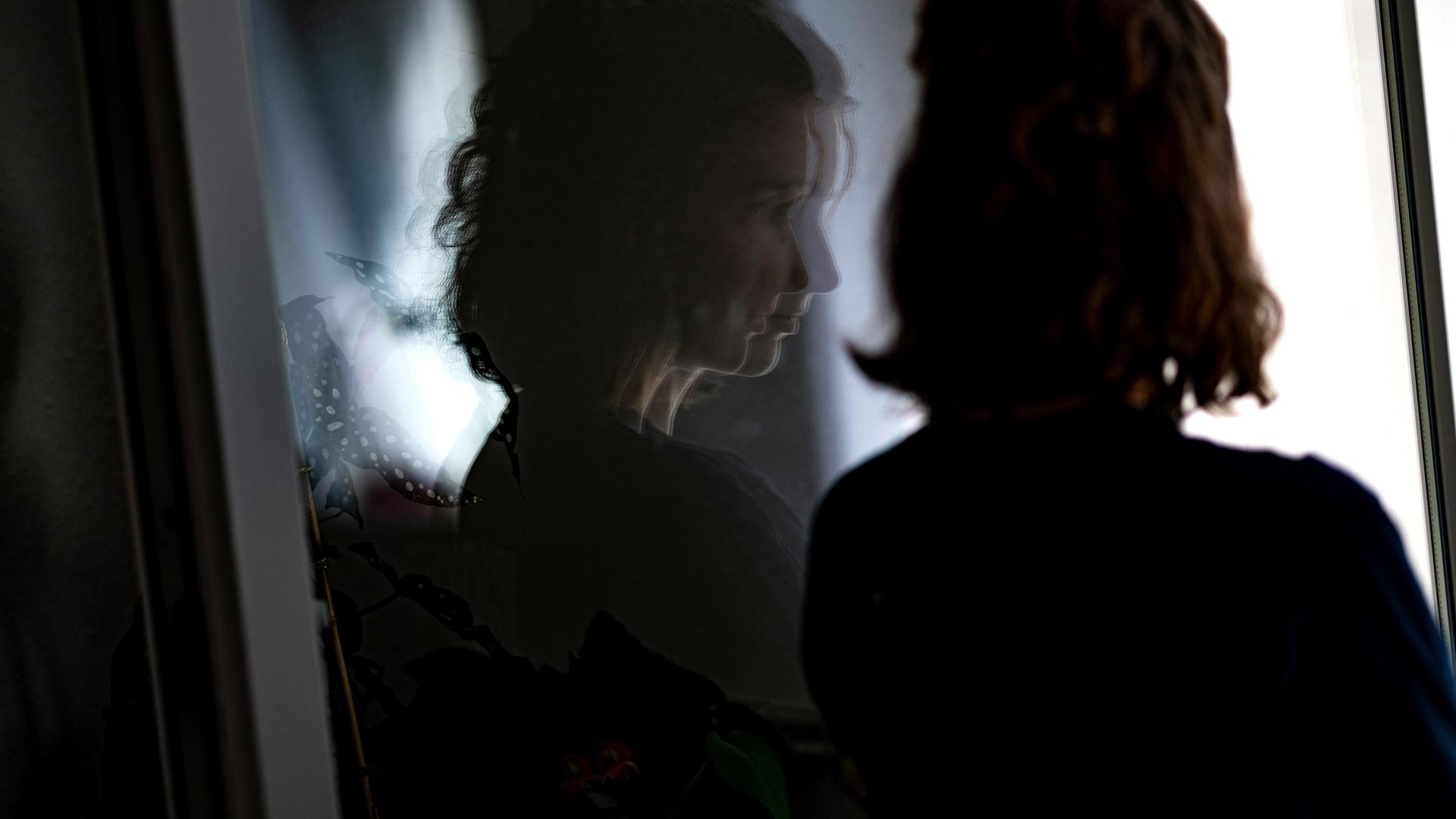 Eine junge Frau steht in ihrer Wohnung an einem Fenster.