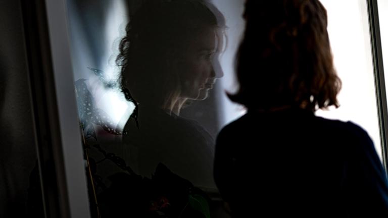 Eine junge Frau steht in ihrer Wohnung an einem Fenster.