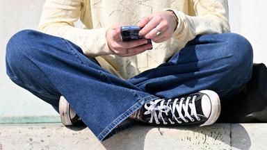 Eine junge Person sitzt im Schneidersitz und hat ein Smartphone in der Hand.