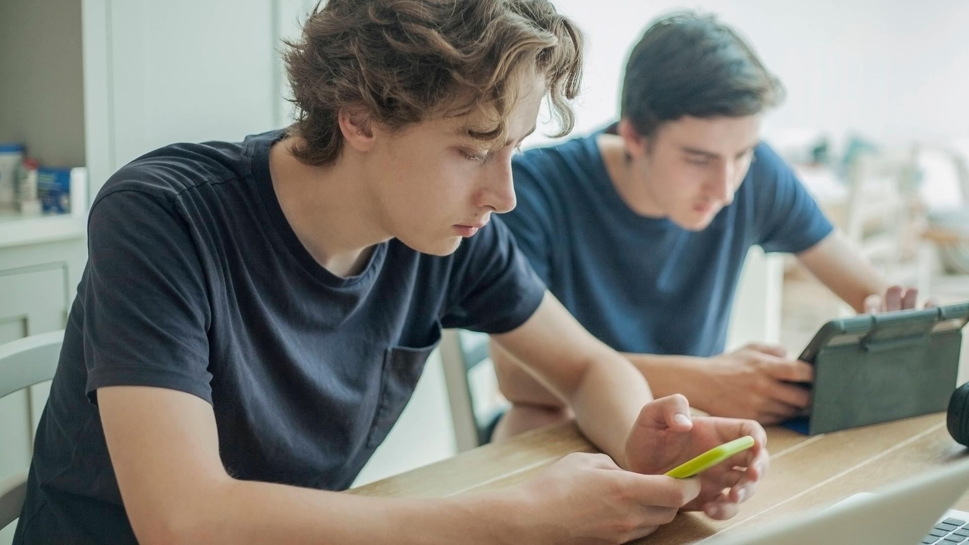 Zwei Jugendliche sitzen an einem Tisch und benutzen ein Smartphone und ein Tablet.