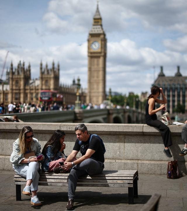 Im Vordergrund sind junge Menschen, die auf einer Bank sitzen. Im Hintergrund ist die Stadt London zu sehen.
