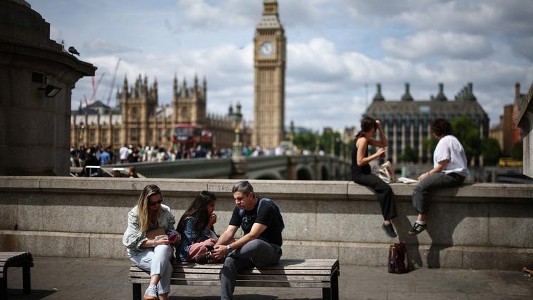 Im Vordergrund sind junge Menschen, die auf einer Bank sitzen. Im Hintergrund ist die Stadt London zu sehen.