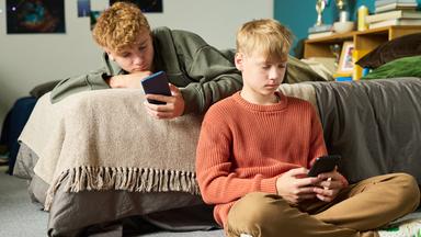 Ein Junge liegt auf einem Bett, ein anderer lehnt daran. Beide schauen auf ihr Smartphone.