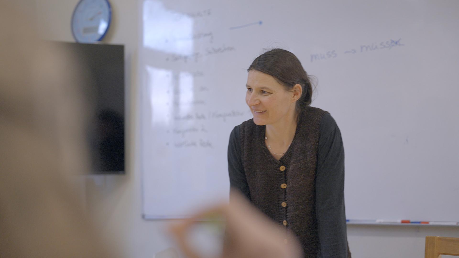 Eine braunhaarige Frau steht in einem Klassenzimmer vor einer Tafel.
