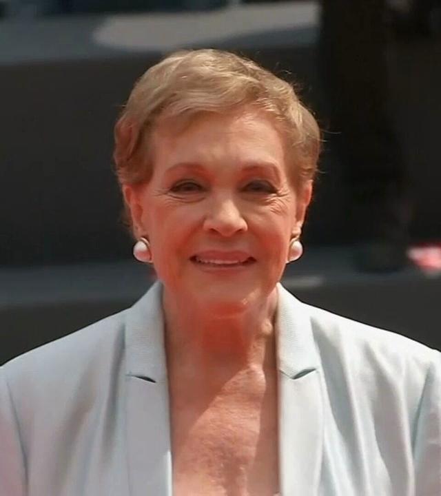 Julie Andrews bei einer öffentlichen Veranstaltung, aufgenommen in einer Nahaufnahme. Sie trägt einen hellen Blazer und runde Ohrringe; der Hintergrund ist unscharf.