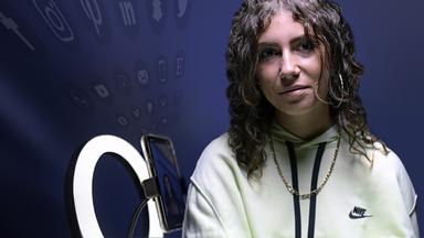 Porträtaufnahme: Laura, eine junge Frau mit Locken und gelbem Pullover, sitzt vor schwarzem Hintergrund und schaut in die Kamera.