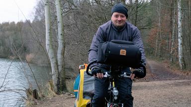 Björn ist mittig im Bild auf seinem Fahrrad zu sehen. Er trägt eine schwarze Mütze und eine graue Jacke. Rechts von ihm sieht man das Ufer eines Sees. Links von ihm führt ein Weg in einen Wald.