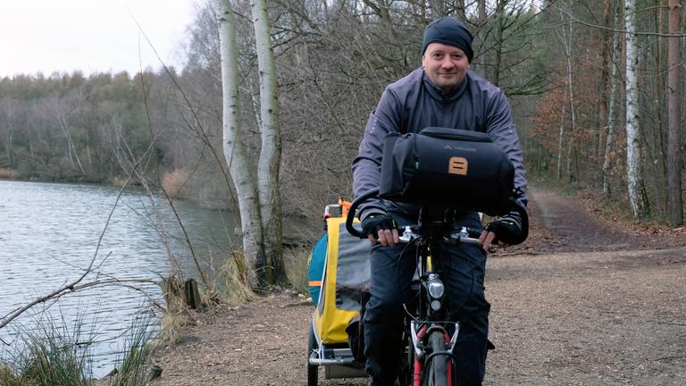 Björn ist mittig im Bild auf seinem Fahrrad zu sehen. Er trägt eine schwarze Mütze und eine graue Jacke. Rechts von ihm sieht man das Ufer eines Sees. Links von ihm führt ein Weg in einen Wald.