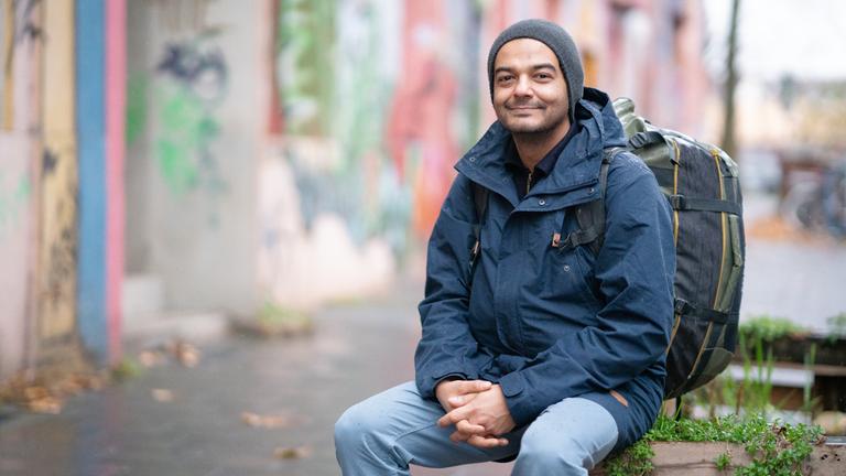 Savan sitzt mit Parka und einem Rucksack auf dem Rücken am Rand eines Bürgersteigs auf einer Mauer und schaut in die Kamera.