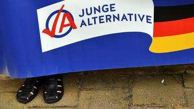 Ein Mann in schwarzen Sandalen hält ein Plakat der AfD-Jugendorganisation Junge Alternative