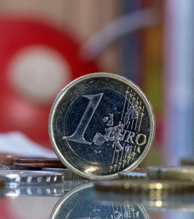 Eine 1 Euro Münze steht auf einem Tisch vor einem Sparschwein.