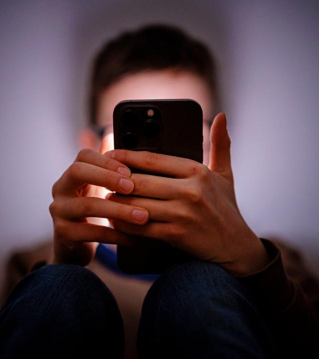 Ein Junge sitzt in einem dunklen Raum an einem Smartphone.
