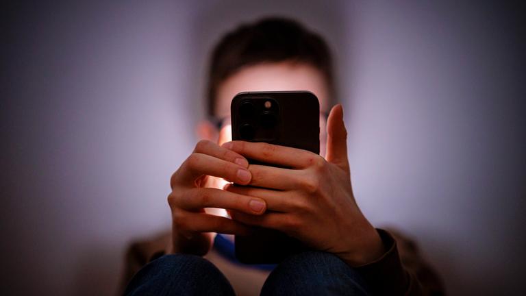 Ein Junge sitzt in einem dunklen Raum an einem Smartphone.