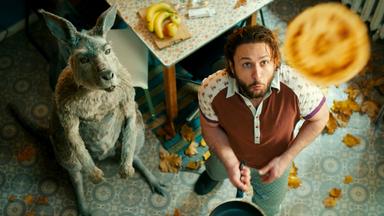 Das Känguru und Marc-Uwe (Dimitrij Schaad) stehen in der Küche und schauen fasziniert zu einem Pfannkuchen hoch, den Marc-Uwe mit der Pfanne in die Luft geworfen hat.