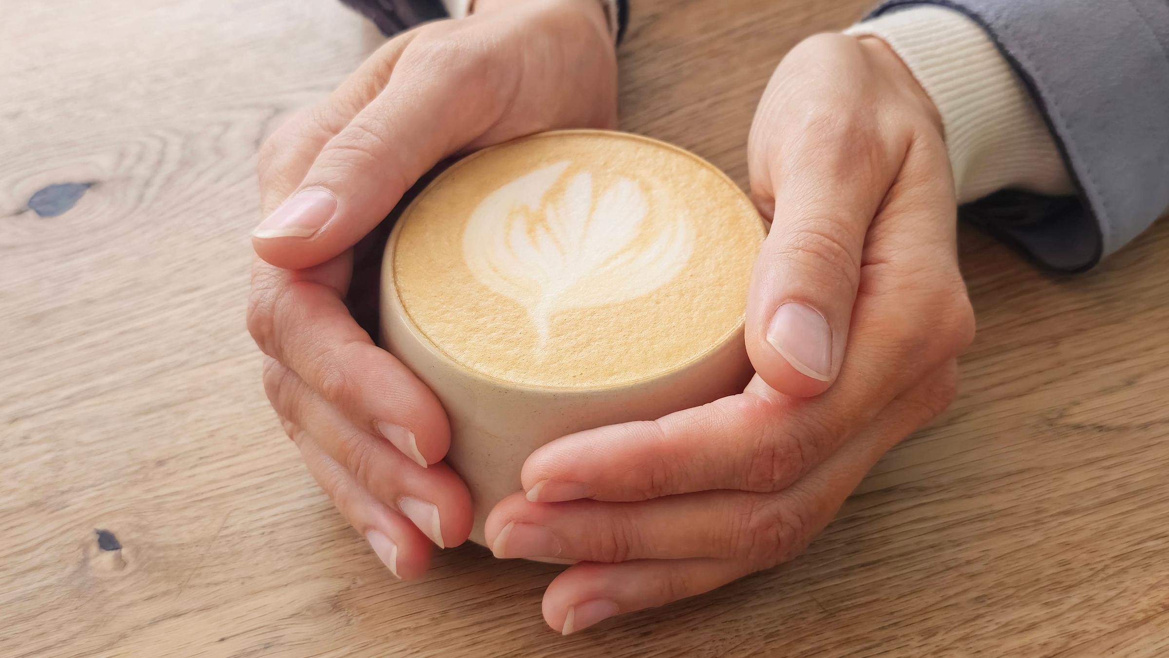Eine Frau hält mit beiden Händen eine auf dem Tisch stehende Tasse mit Milchkaffee.