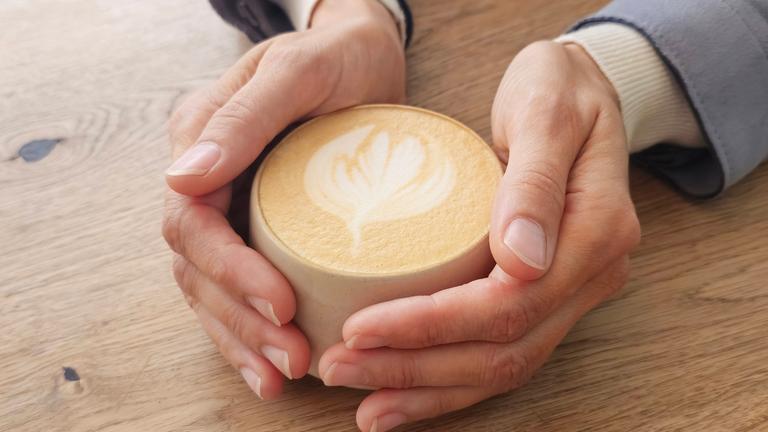 Eine Frau hält mit beiden Händen eine auf dem Tisch stehende Tasse mit Milchkaffee.