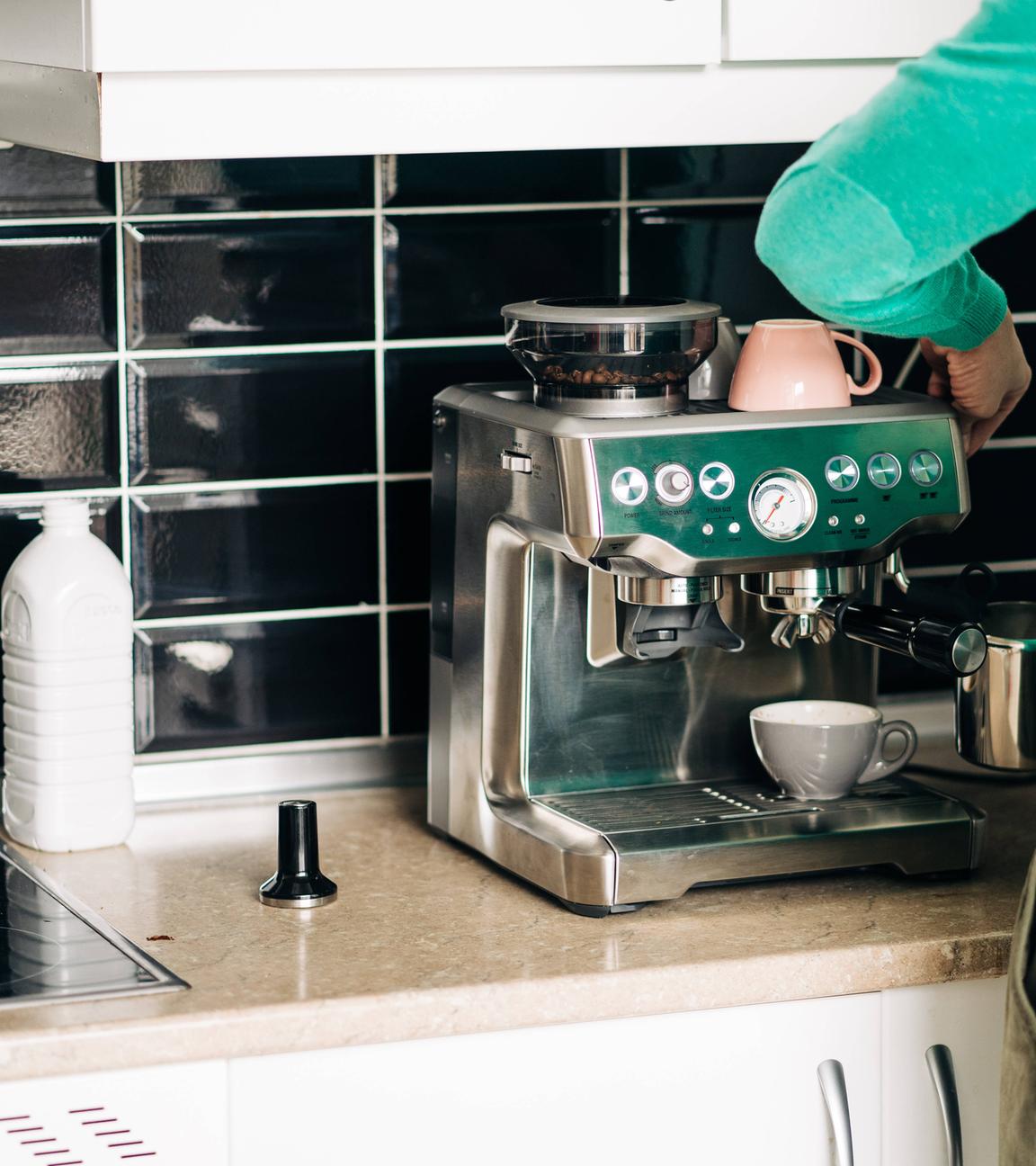 Mann steht vor einer Espressomaschine und reinigt sie. Neben der Maschine steht eine Flasche mit Reinigungsmittel.