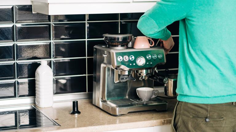 Mann steht vor einer Espressomaschine und reinigt sie. Neben der Maschine steht eine Flasche mit Reinigungsmittel.