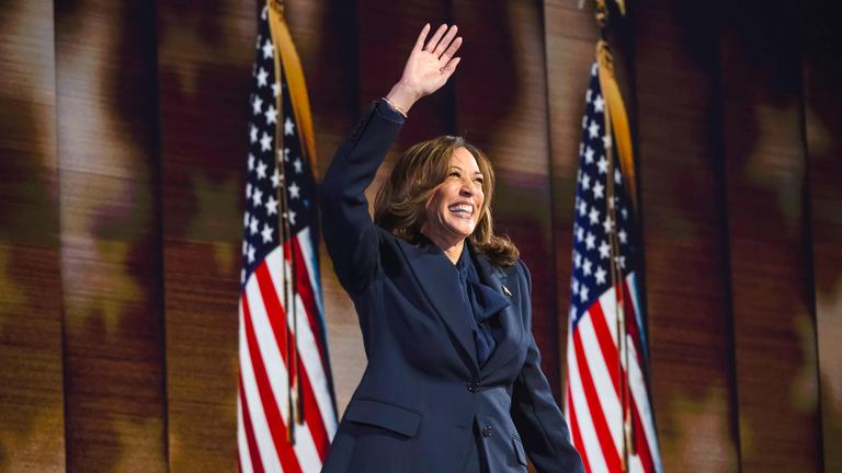 Eine Frau mittleren Alters mit langen braunen Haaren und gekleidet in einem Businessanzug steht zwischen zwei US-amerikanischen Flaggen und hebt lachend eine Hand hoch über den Kopf.