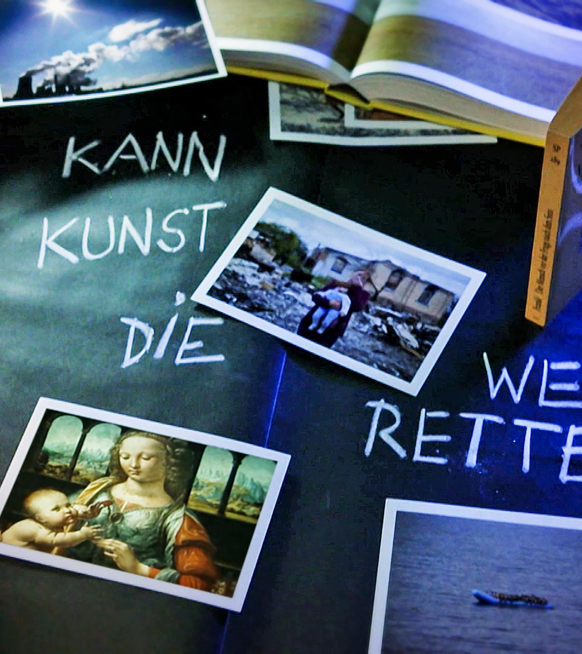 Auf einem Tisch sind viele Fotos verteilt, dazwischen steht mit Kreide geschrieben: Kann Kunst die Welt retten?