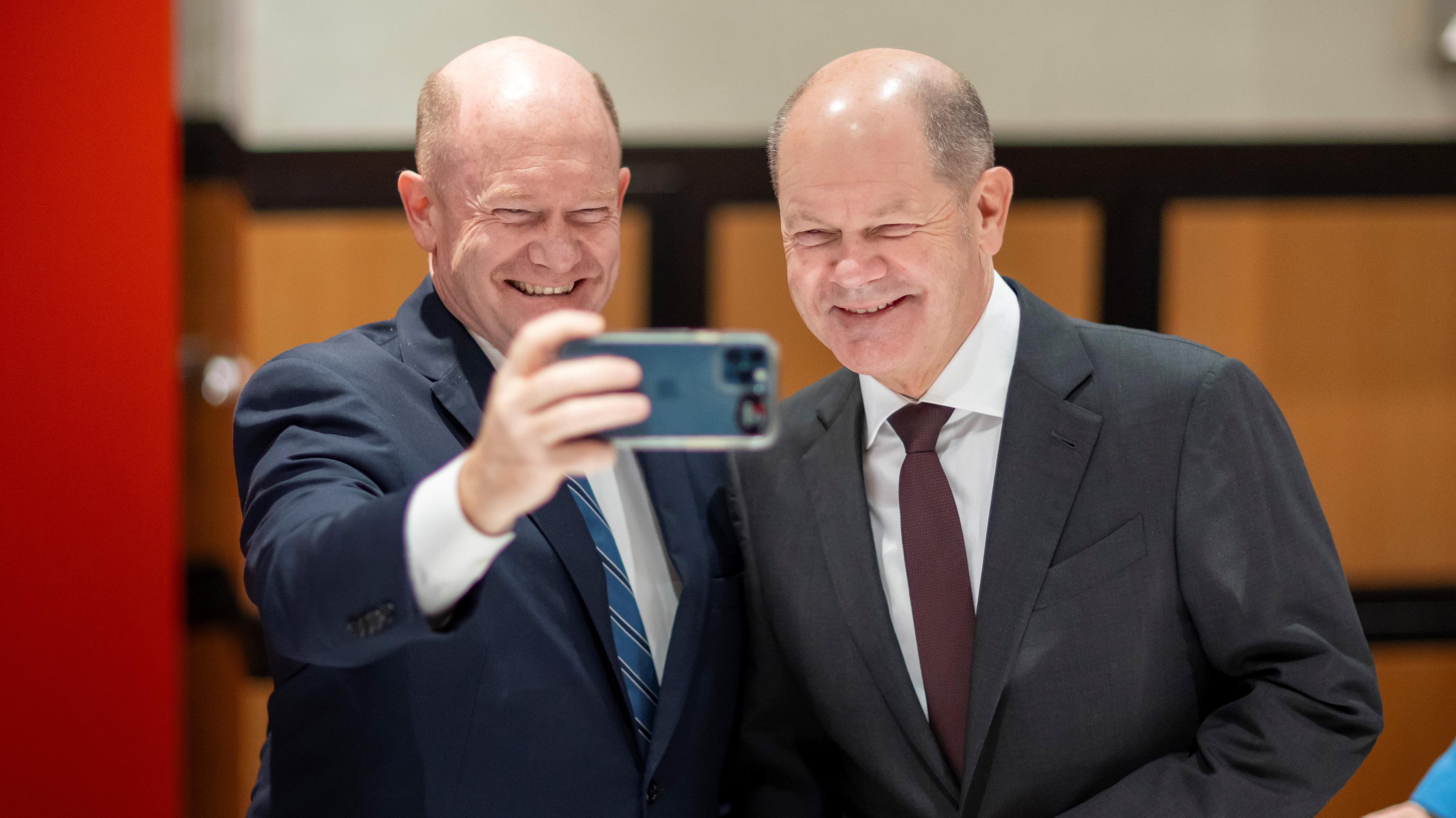 Rechts ist Kanzler Scholz zu sehen. Zusammen mit Chris Coons, der links steht, macht er ein Selfie.