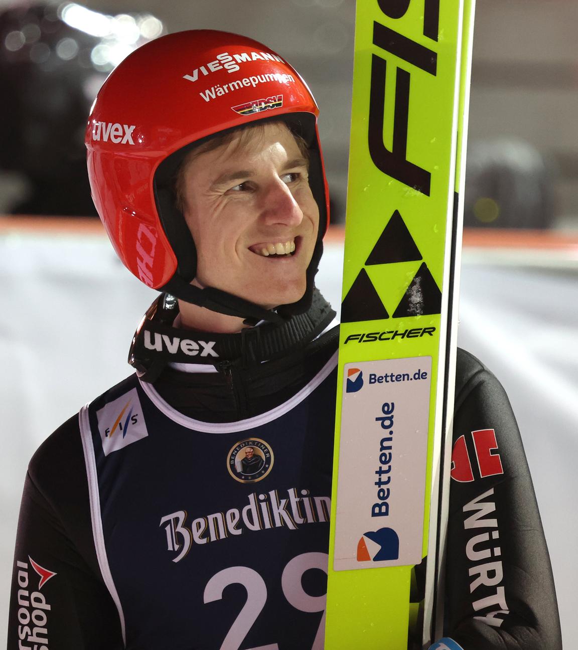 Karl Geiger lachend in Skisprunguniform und den Skiern neben sich aufgestellt