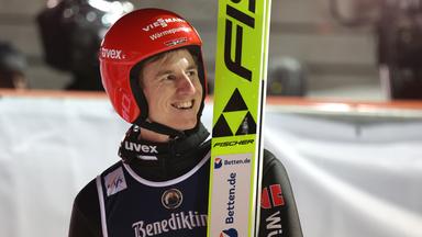 Karl Geiger lachend in Skisprunguniform und den Skiern neben sich aufgestellt