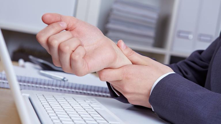 Eine Hand stützt das Handgelenk der anderen Hand über einem Laptop. Daneben liegt ein Ringblock mit einem Kugelschreiber.