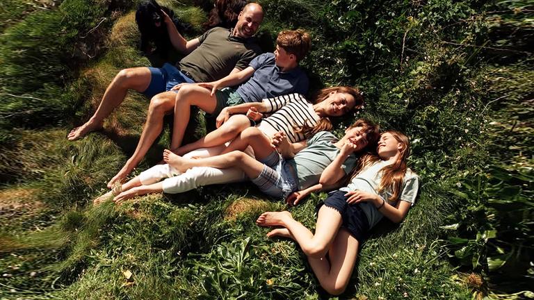 Ein Bild von Kate und William, wie sie mit ihren drei Kindern auf einer Grasfläche liegen und lächeln.