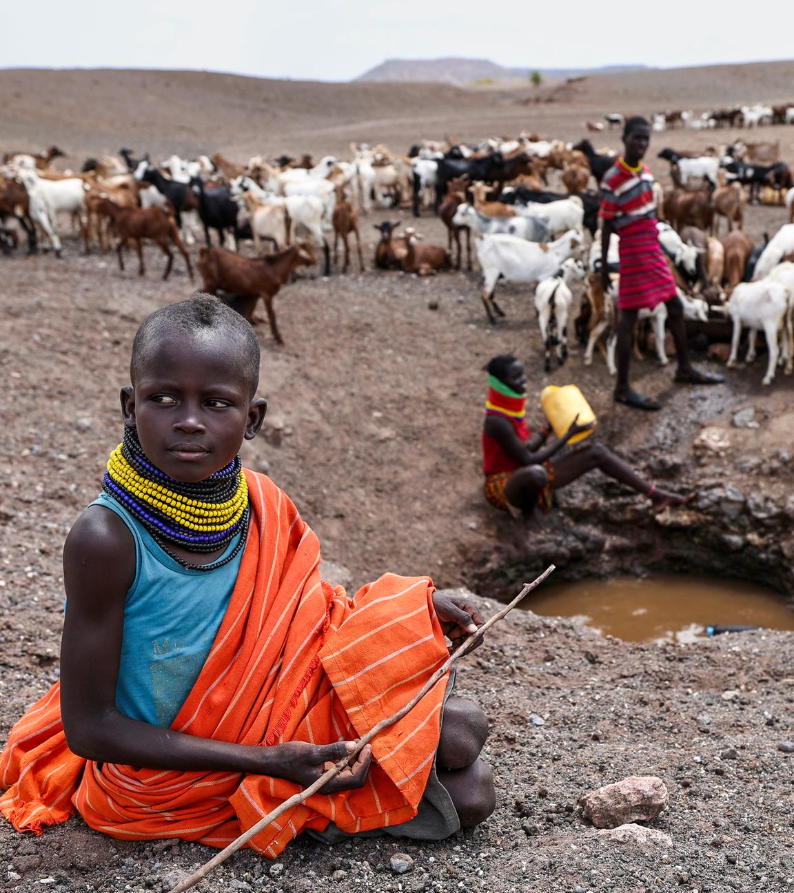 Ein Mädchen sitzt auf dem Boden und hält einen Stock in der Hand. Im Hintergrund sind weitere Personen und eine Herde Ziegen.