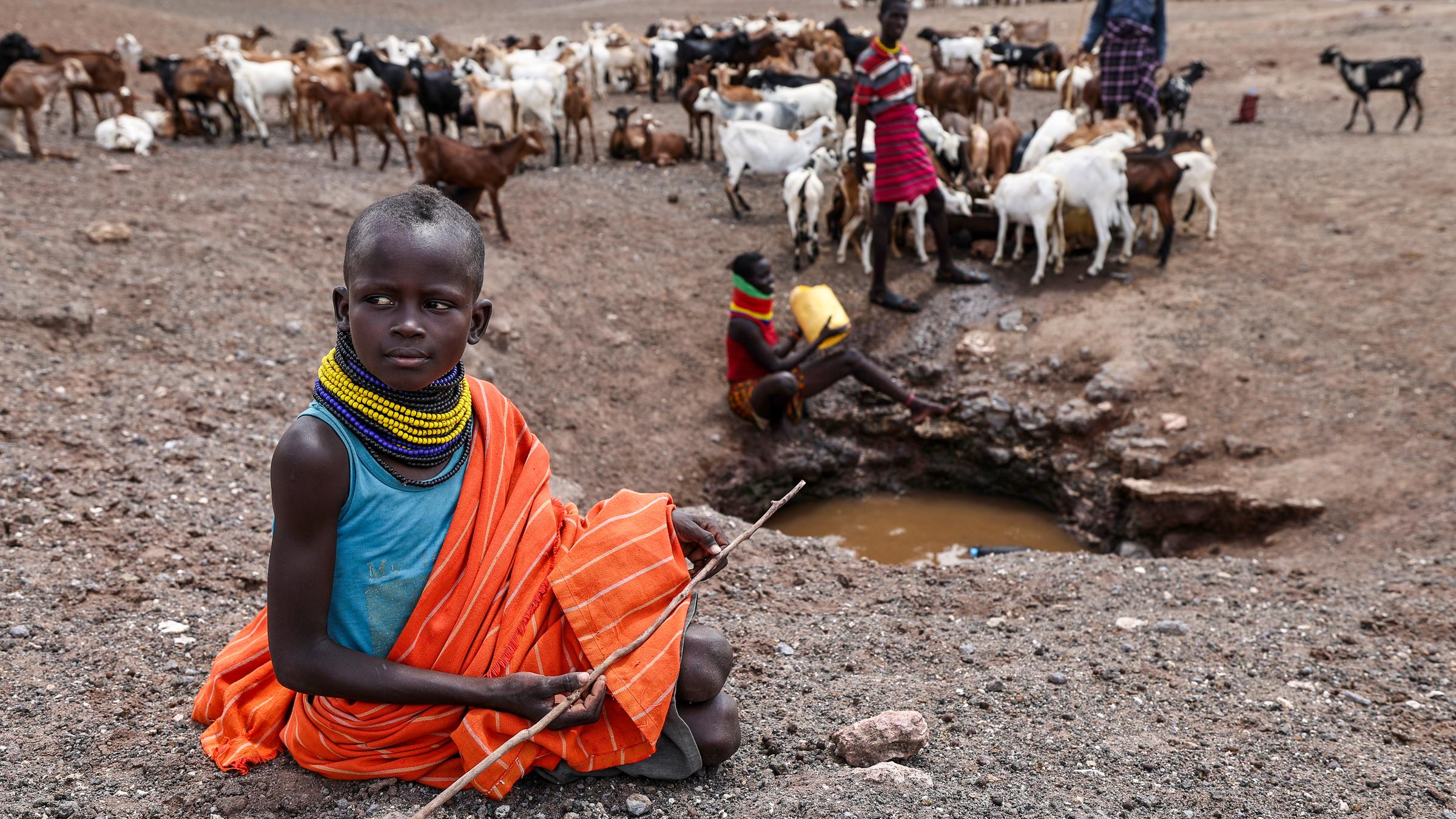 Ein Mädchen sitzt auf dem Boden und hält einen Stock in der Hand. Im Hintergrund sind weitere Personen und eine Herde Ziegen.