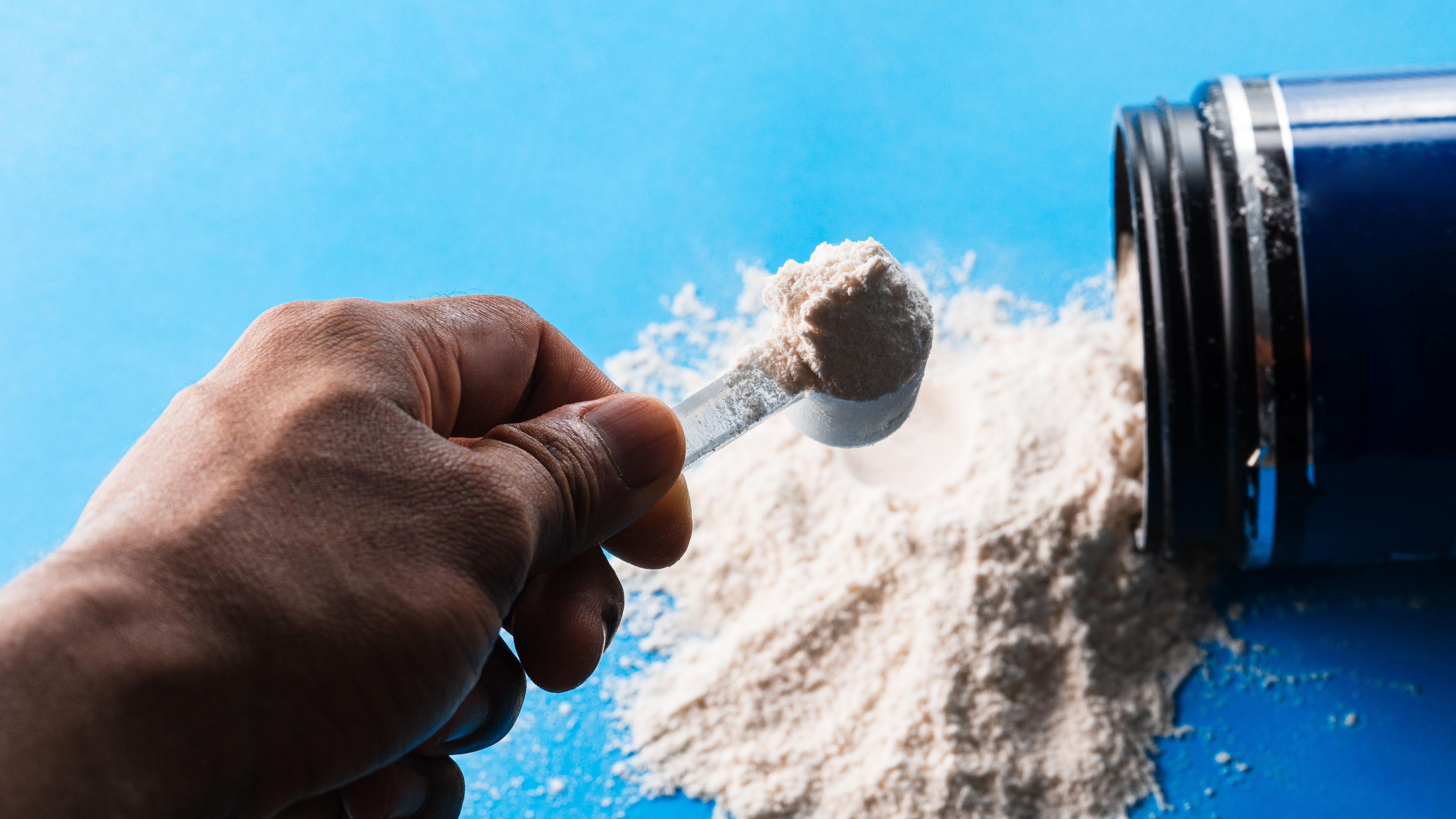 Hand hält einen Portionierlöffel voller weißem Pulver Keratin. Daben liegt eine geöffnete Dose.