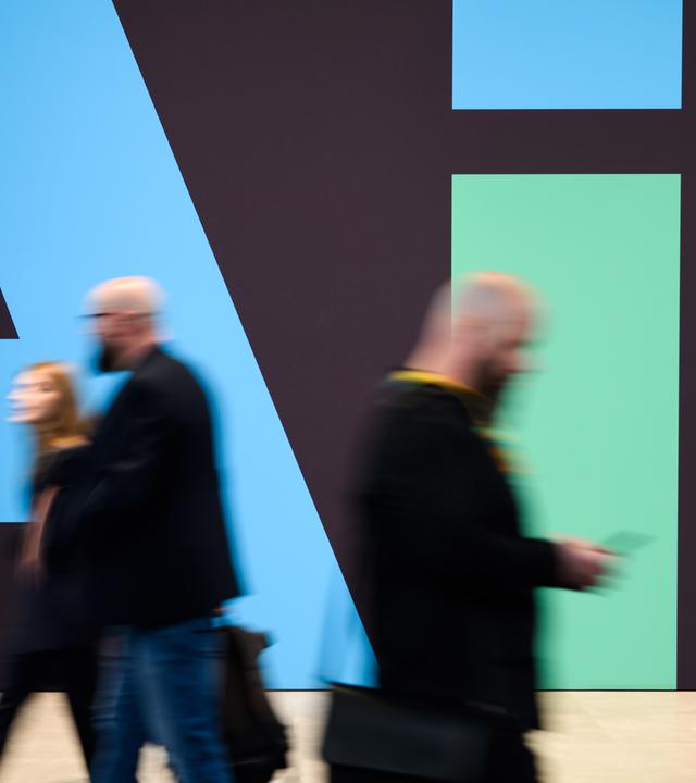 Das Bild zeigt Gäste der Hannover-Messe, die an einem Stand von Siemens mit einem "AI"-Schriftzug vorbeigehen.