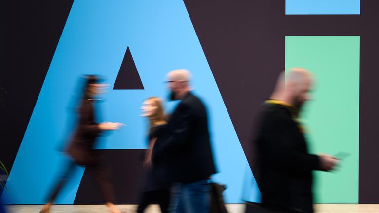 Das Bild zeigt Gäste der Hannover-Messe, die an einem Stand von Siemens mit einem "AI"-Schriftzug vorbeigehen.