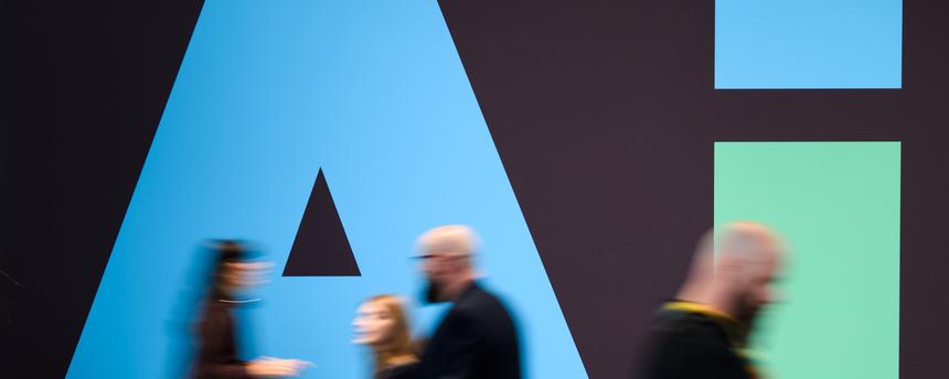 Das Bild zeigt Gäste der Hannover-Messe, die an einem Stand von Siemens mit einem "AI"-Schriftzug vorbeigehen.