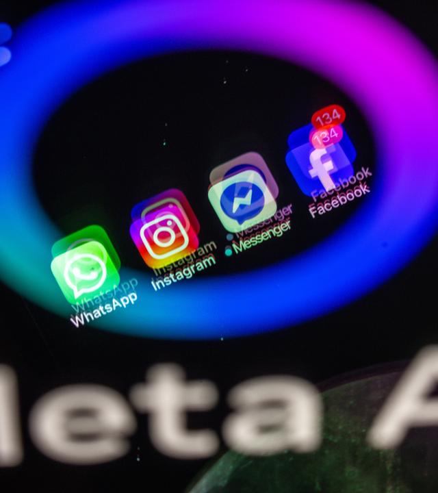 Symbolbild: Die Apps Facebook, Messenger, Instagram und WhatsApp werden auf einem Smartphone-Display angezeigt in dem das Logo der KI-Anwendung Meta AI spiegelt.