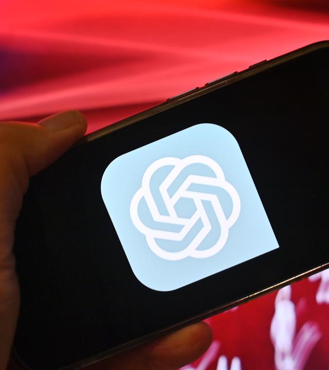 Hand zeigt ein Smartphone mit dem ChatGPT Logo vor rotem Hintergrund