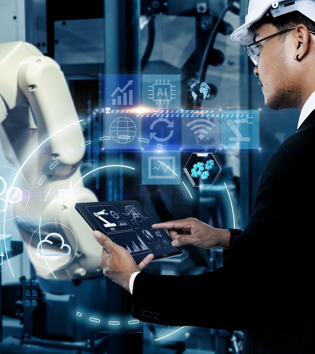 In einer Industrieanlage steht rechts ein Mann mit weißem Schutzhelm und Brille. Er hält ein Tablet und berührt dessen Bildschirm. Links neben ihm ist ein heller Roboterarm zu sehen. Blaue, über das Bild gelegte Grafiken zeigen unter anderem ein KI-Symbol, Diagramme und vernetzte Zahnräder.