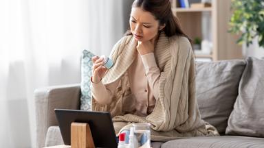 Eine Frau sitzt mit einer Decke um die Schultern auf einem Sofa und schaut auf ein Tablet, das vor ihr steht.